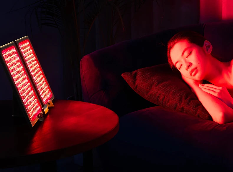 A red light therapy panel glowing with red light sits on a small table next to a woman sleeping on a couch in a dimly lit room, illuminated by the red light.