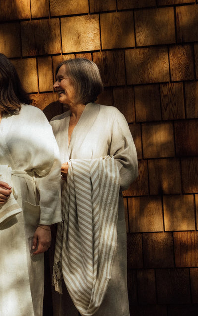 Two people wearing cream-colored robes standing against a warm wood shingle wall, with one person holding a striped cream and gray blanket.