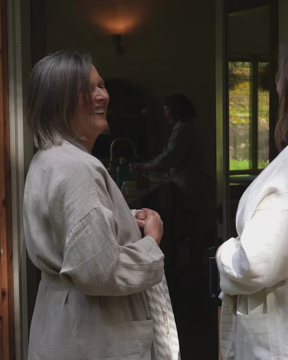 A woman with gray hair wearing a light beige linen robe stands in a doorway, smiling and looking to the side. Another person in a white robe is partially visible on the right, and a softly lit kitchen area is visible in the background.