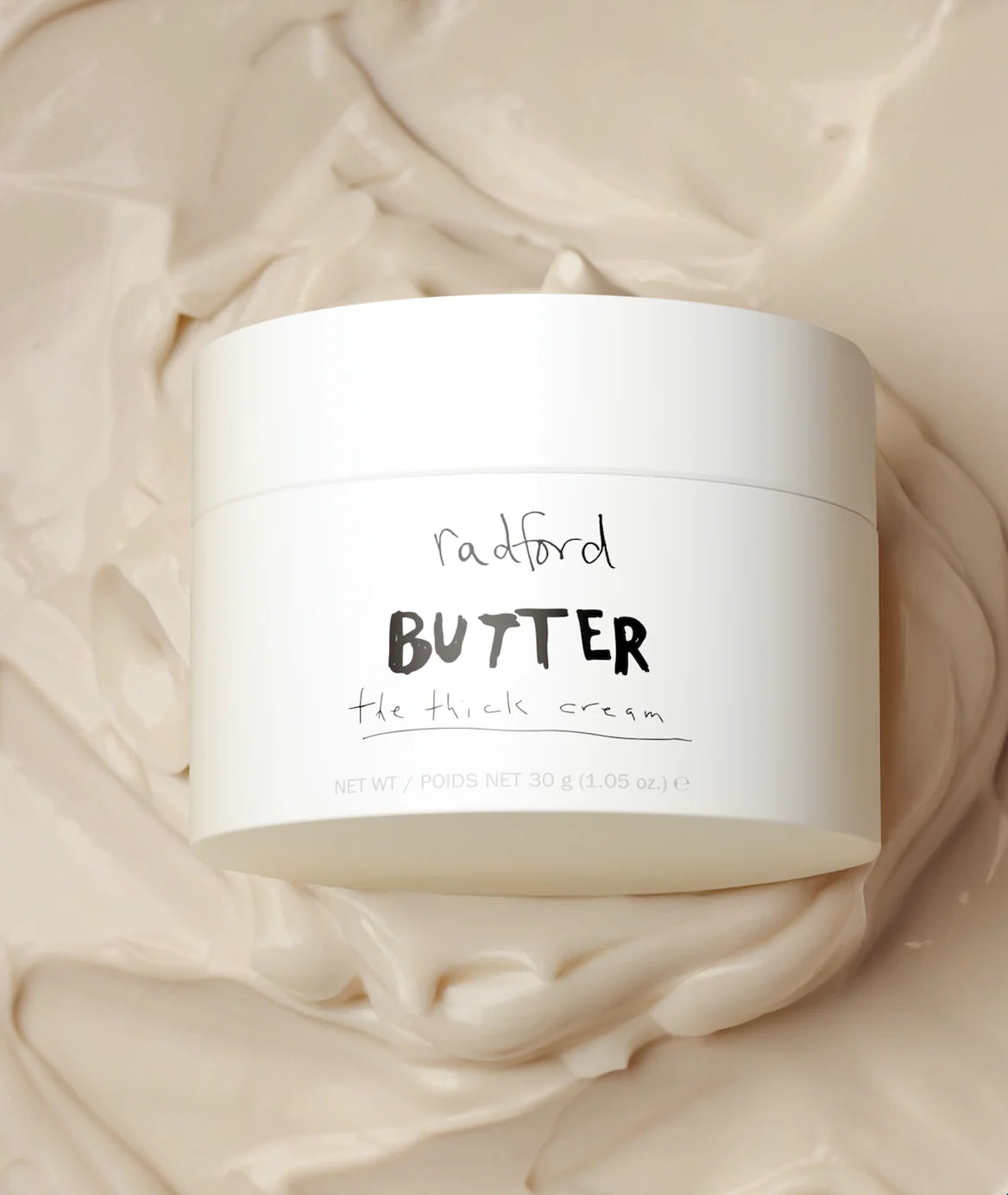 A white jar of Radford Butter The Thick Cream resting on a background of smooth, swirled cream-colored product.