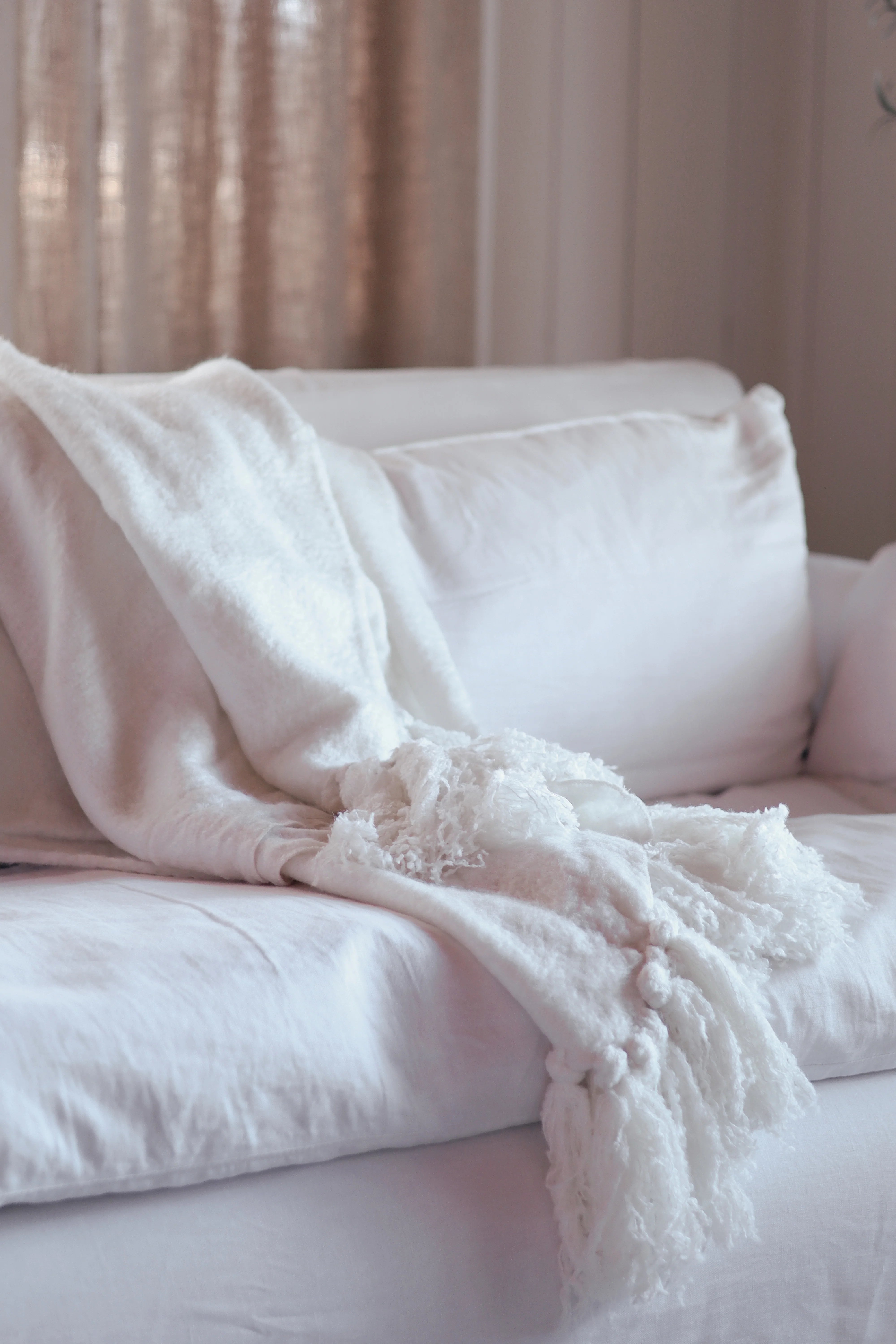 A soft white fringed throw blanket draped over a white sofa with pillows in a softly lit bedroom setting.