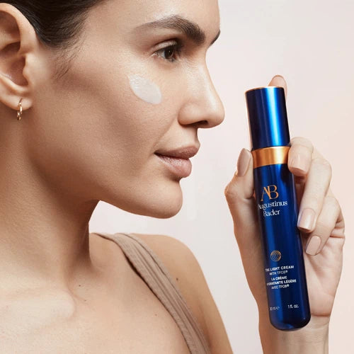A woman in profile holds a blue and gold AB Rhythm Blacker skincare bottle next to her cheek, which has a white cream smear on it, against a soft beige background.