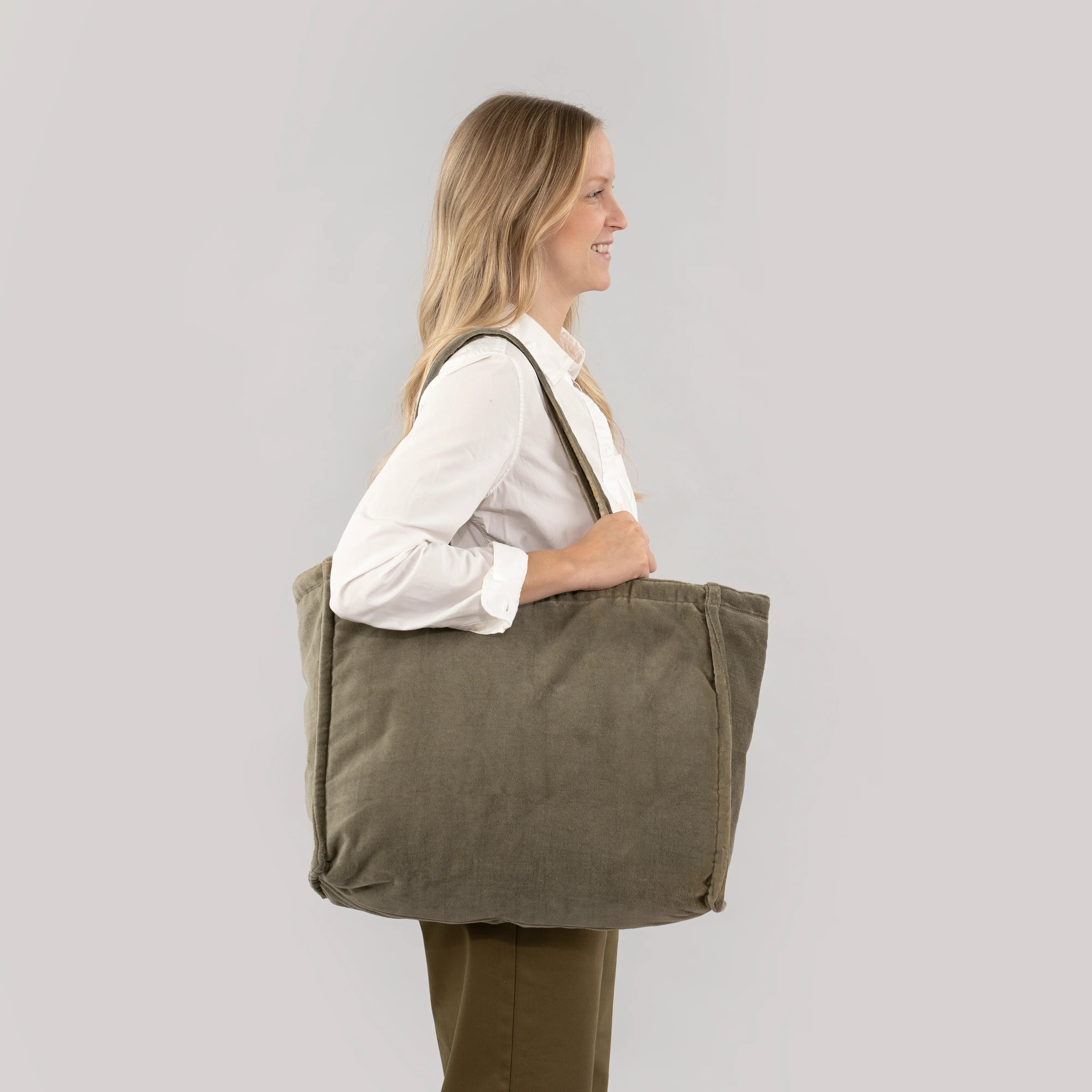 A woman in a white shirt and olive pants carries a large olive green canvas tote bag with a shoulder strap, shown from the side against a light gray studio background.