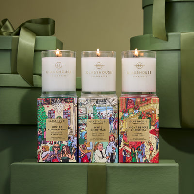 Three lit glasshouse fragrances candles in illustrated boxes with green gift boxes in the background.
