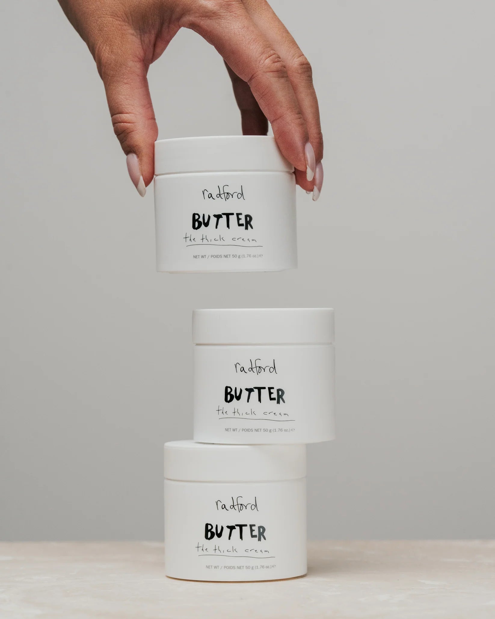 Three white jars of 'naughty BUTTER' hand cream stacked vertically, with a hand holding the top jar against a neutral gray background.