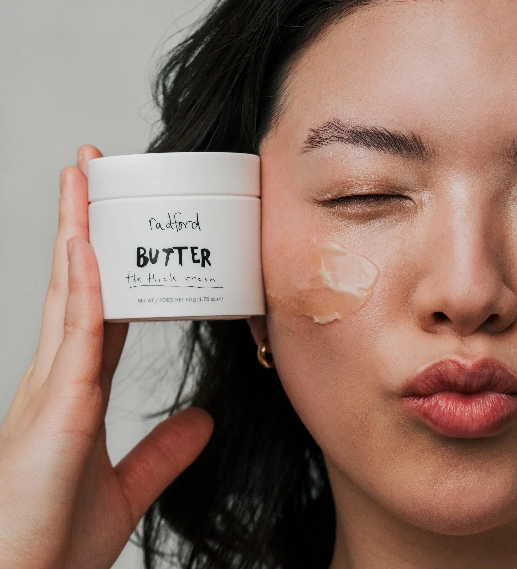 A close-up of a woman holding a white jar of Radiford Butter face cream next to her face, which has a smear of the cream on one eye area.