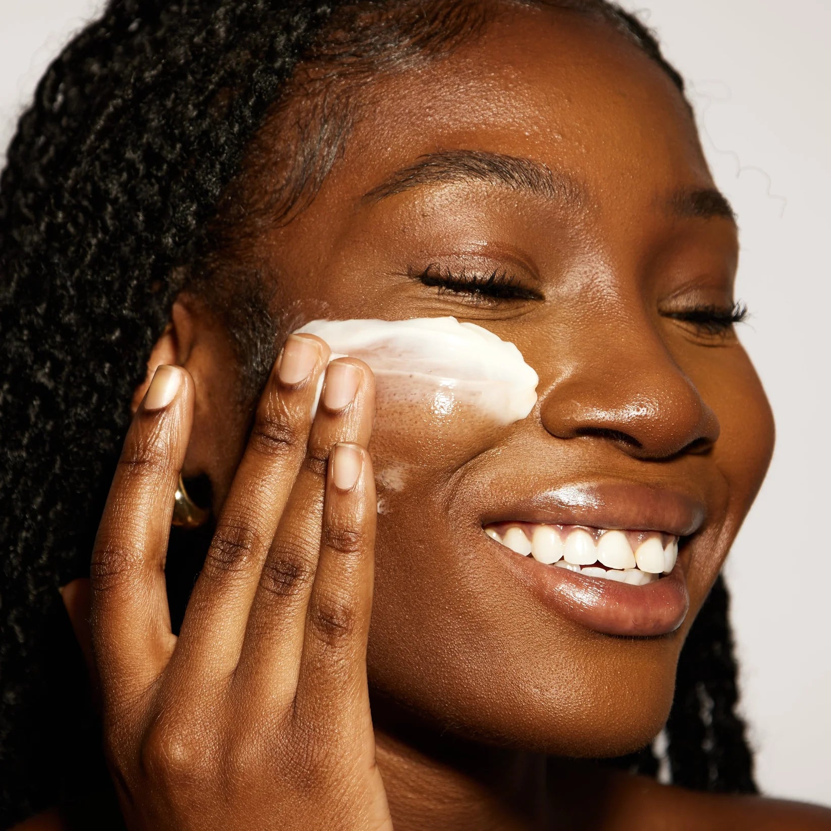A close-up of a woman with dark curly hair applying a thick white cream or balm to her cheek with her fingers against a plain light background.