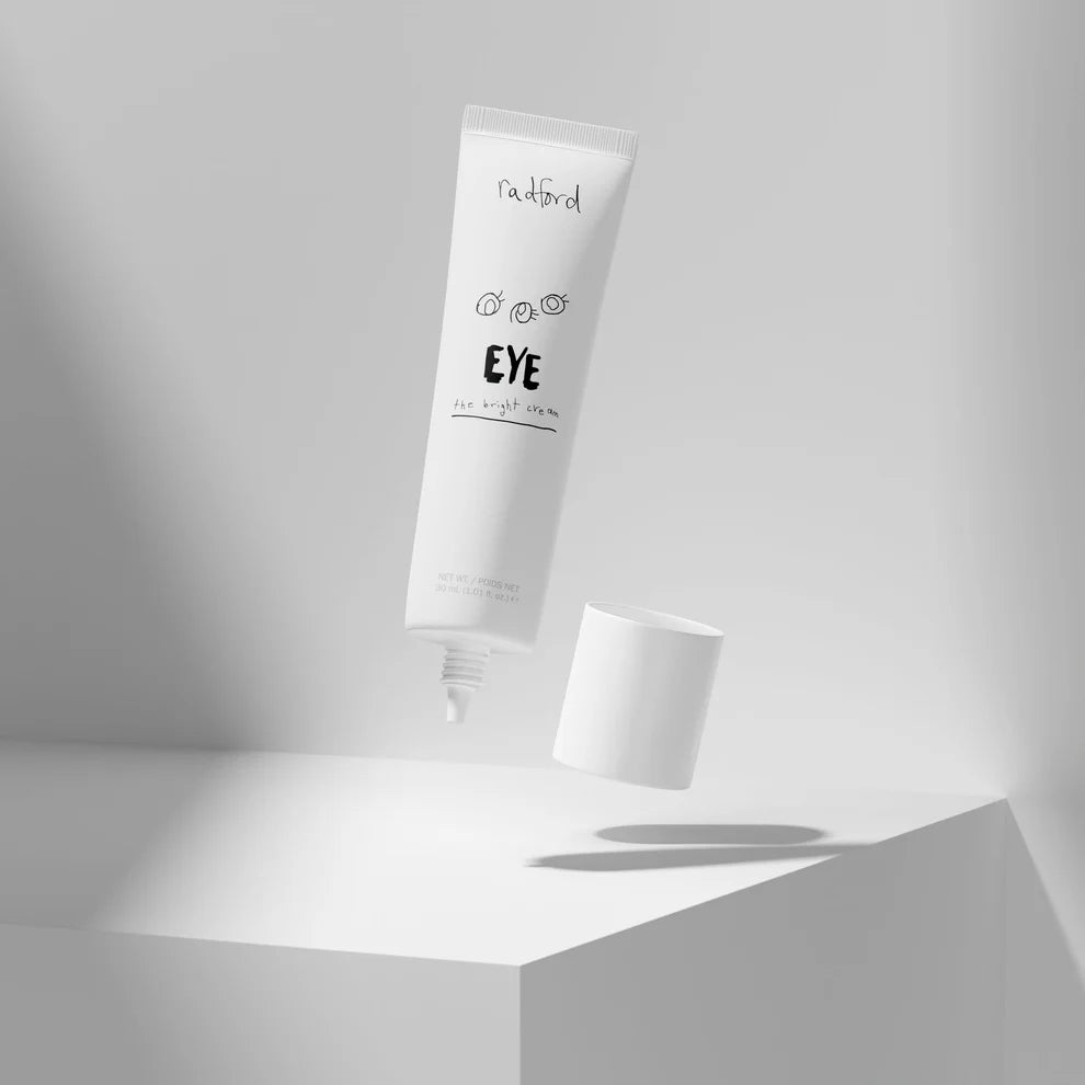 A white cosmetic tube labeled 'naked' and 'EYE' floating above a white geometric pedestal, with its cap removed and a small amount of cream dripping from the tube tip in a clean studio setting.