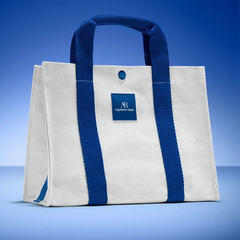 A white canvas tote bag with thick blue webbed handles and side straps, featuring a blue brand patch on the front that reads 'AB Agricultural Banks'. The bag is photographed on a blue gradient studio background.